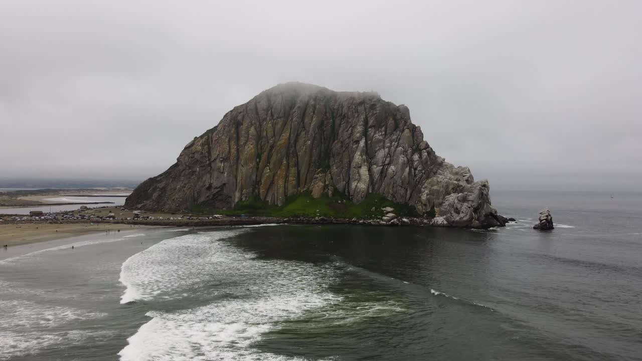 Aerial zoom out shot of waves rolling onto the beach and Morro Rock in coastal California. The rock is slightly obscured by the fog that has rolled in from sea as the scene is revealed.