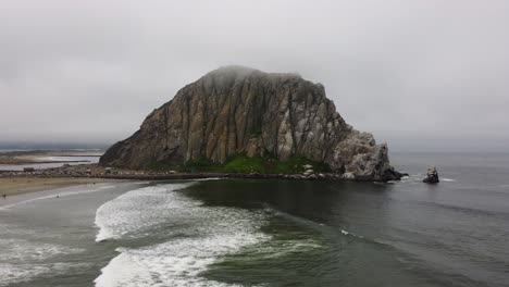 aerial zoom out shot of waves rolling onto the beach and morro rock in coastal california. the rock is slightly obscured by the fog that has rolled in from sea as the scene is revealed.