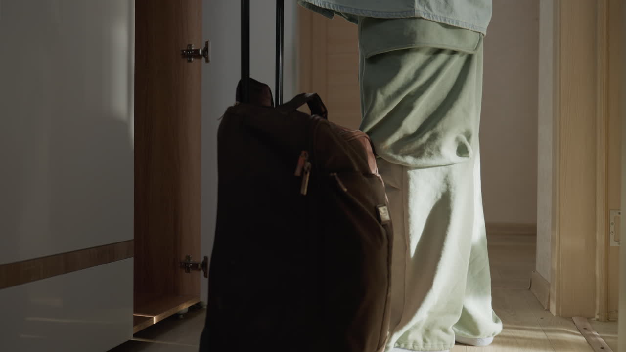 Mujer entrando en un estrecho pasillo de apartamento con una maleta, tirando de una maleta con ruedas hacia el armario, suave luz matutina proyectando largas sombras, ambiente tranquilo, nueva inquilina llegando, pasos tentativos hacia una puerta abierta