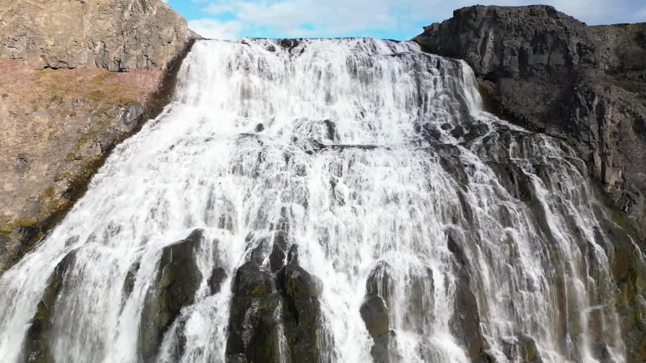 Splendidi clip video aerei della maestosa cascata di Dynjandi nei fiordi occidentali dell'Islanda, che mostrano potenti acque bianche che scendono a cascata su rocce vulcaniche scure sotto un cielo blu