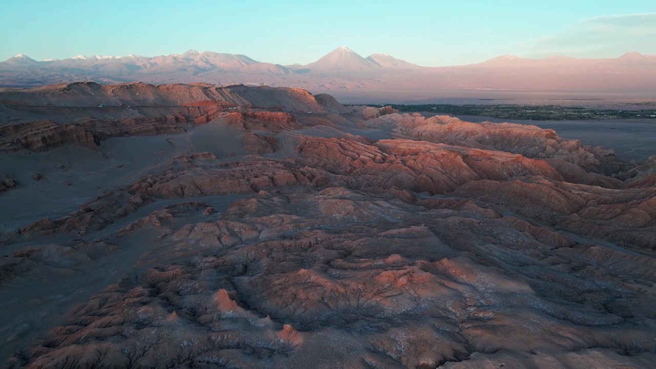 Volo in discesa sopra le formazioni rocciose texturizzate della Valle della Luna al tramonto, che rivela una città oasi, una distesa di sale e cime vulcaniche andine innevate in un cielo crepuscolare rosa. San Pedro de Atacama, Cile.