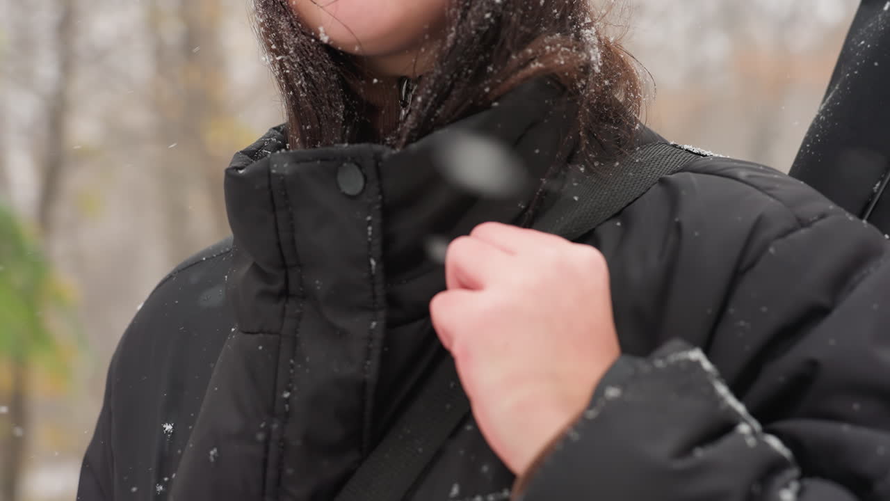 옷과 머리카락에 눈알이 있는 검은색 겨울 재 줄을 조정하는 여자의 클로즈업, 추운 날씨를 강조하고, 배경에는 눈 인 공원에서 흐릿한 가을 나무가 있습니다.