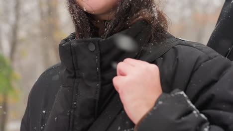 close-up of lady adjusting black winter jacket strap with snowflakes on clothing and hair, highlighting cold weather, background features blurry autumn trees in snow-covered park