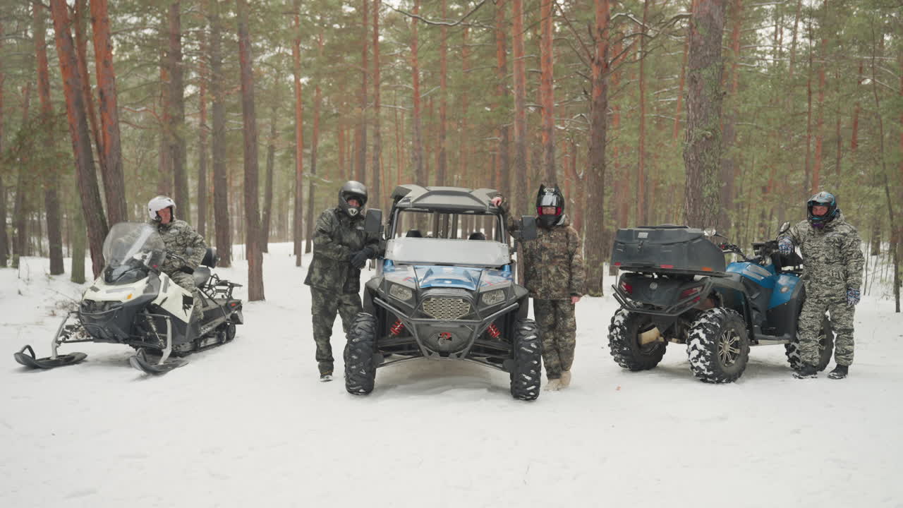 Schieramento di ATV e motoslitte nella foresta innevata. Tre piloti con caschi e tute isolate preparano l'equipaggiamento da spedizione. La guida controlla il verricello e il meccanico ispeziona le sospensioni, mentre i fiocchi di neve cadono sotto l'alta chioma dei pini.