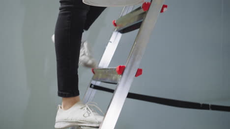 lady goes up step ladder holding paint bucket in room