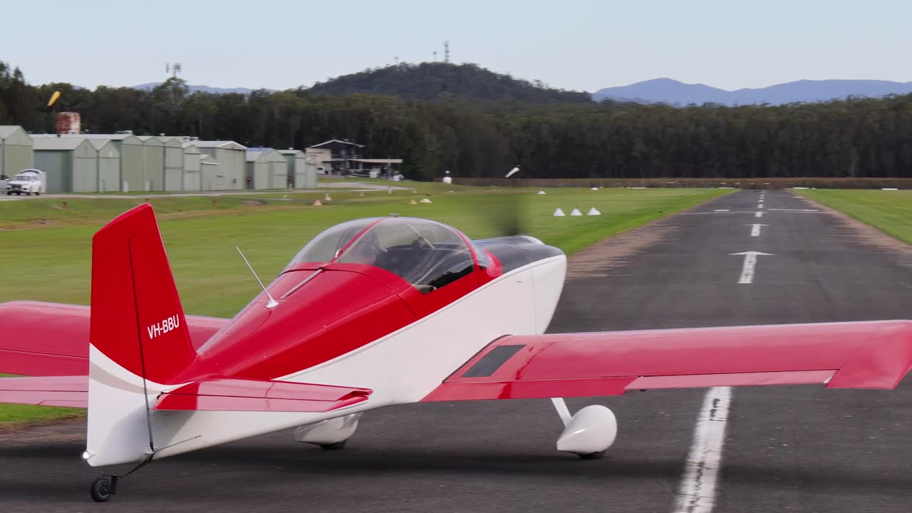 A small red aircraft takes off from a runway with mountains in the background.