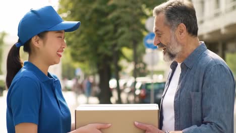 a delivery person wearing a blue uniform smiles as they hand a cardboard package to a man.