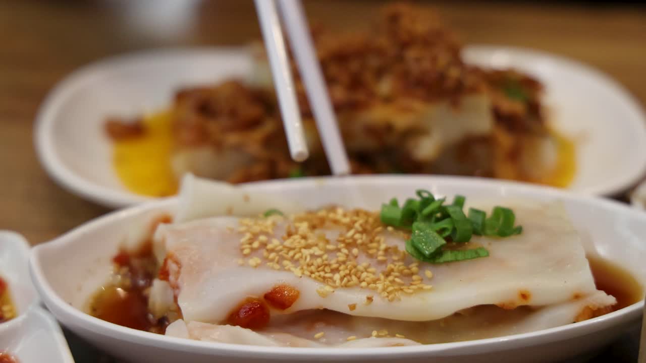 Chopsticks grasp and lift a sesame-topped steamed rice noodle roll from a plate, with warm indoor lighting and shallow depth of field highlighting the food