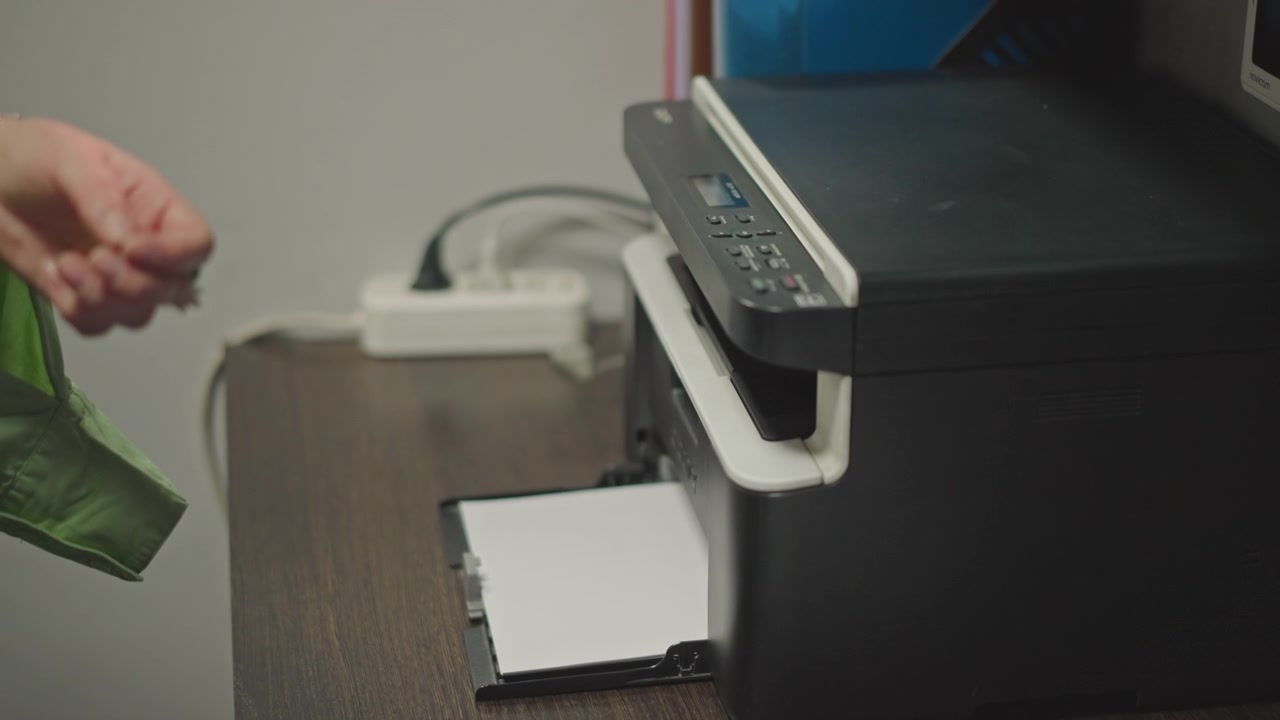 Music teacher prints sheet music while retrieving fresh pages from compact office printer, closeup on hand guiding paper tray, soft task lighting on tidy desk, visible cables and supplies, calm