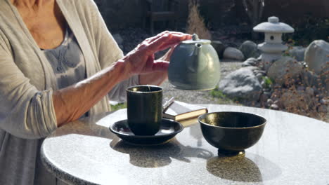 a woman placing an herbal tea bag into a cup and pouring hot water from a teapot then letting it steep