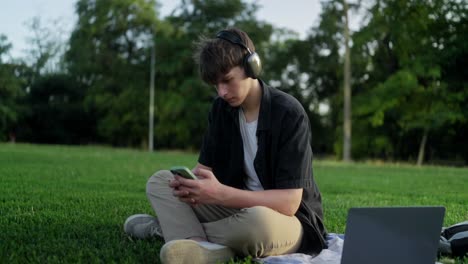 hombre sentado en la hierba en un parque con auriculares, mirando su teléfono
