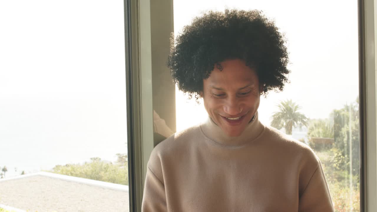 African American man scrolling smartphone and smiling while glancing out glass window at hillside
