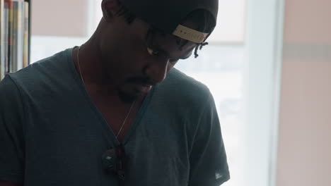 close up of young man in dark cap and grey t-shirt looking down while adjusting glasses hanging from necklace in bright library setting, appearing focused and thoughtful in quiet moment