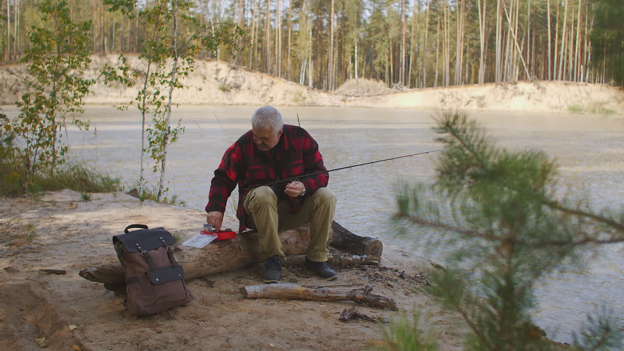 fisherman is preparing baits and lures for spin fishing choosing from box holding rod in hand sitting on coast of lake in forest