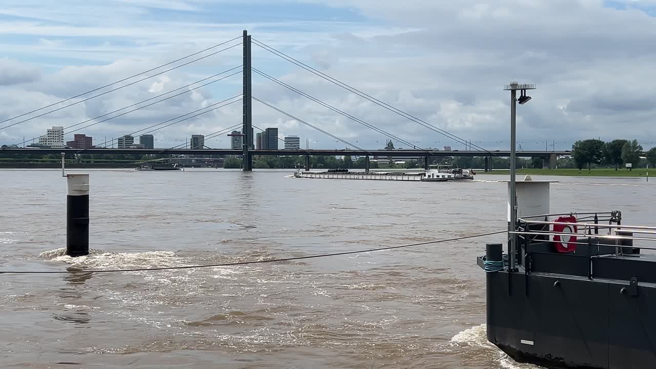 Traffico fluviale sul Reno a Düsseldorf con nave cargo, ponte moderno e skyline della città. Scena urbana sul lungofiume con acqua che scorre sotto un cielo nuvoloso
