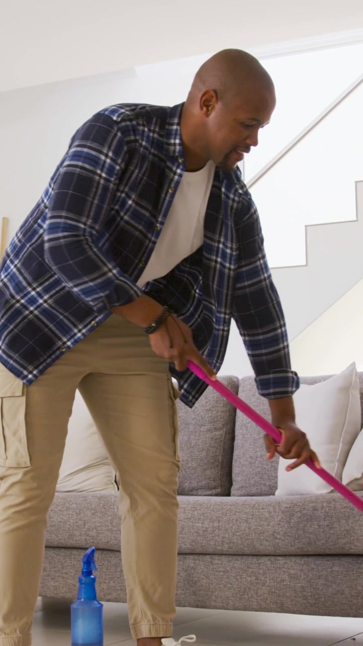 Vertikal video: Strekker seg etter en blå sprayflaske, noe som får en mann i rutete skjorte til å moppe gulvet ved sofaen med en rosa mopp.