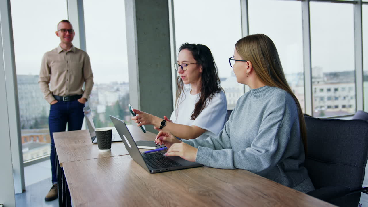 Blonde and brunette females sit at the desk in front of laptop. Male colleague standing at the window addresses to ladies.