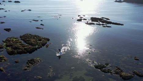 órbita aérea de surf de remo en agua clara a través de rocas cerca de una playa con el sol reflejado en el fondo en el atlántico