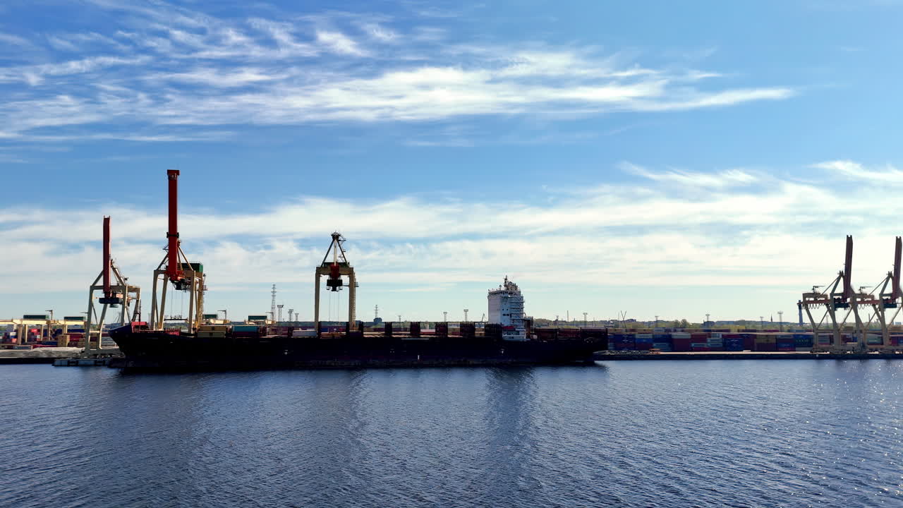 Massive cargo vessel is docked at an industrial shipping port, flanked by towering cranes and stacks of containers under a bright blue sky with scattered clouds