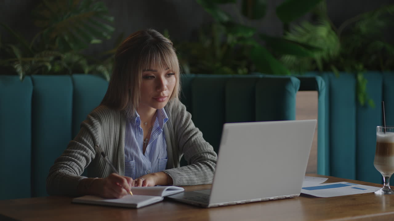 Asian woman working on her laptop and writing in his notebook sitting at a table. Working in coffee shop. woman looking to a laptop screen and making notes in her notebook. studying online.