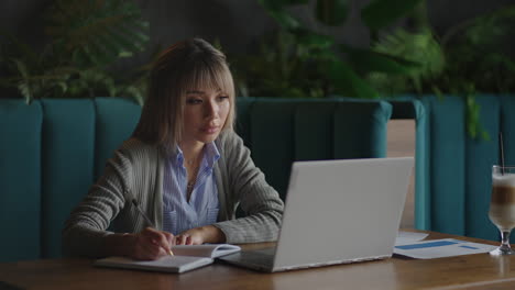 Asiatische-Frau,-Die-An-Ihrem-Laptop-Arbeitet-Und-In-Sein-Notizbuch-Schreibt,-Sitzt-An-Einem-Tisch.-Arbeit-Im-Café.-Frau-Schaut-Auf-Den-Bildschirm-Ihres-Laptops-Und-Macht-Sich-Notizen-In-Ihrem-Notizbuch.-Online-Studieren.