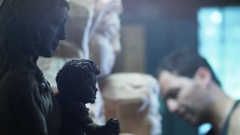 adult sculptor works intently in workshop, flanked by a detailed clay maquette and a larger wooden model of the virgin mary and child, both carefully crafted and aligned in his creative process