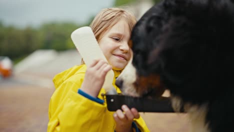 primer plano una adolescente con una chaqueta amarilla le da agua a un perro negro de un cuenco especial para beber mientras camina con su mascota en el parque después de la lluvia