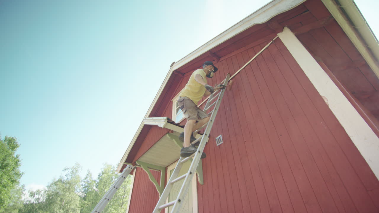 Man on ladder brushing off peeling paint from falu red house, upwards view