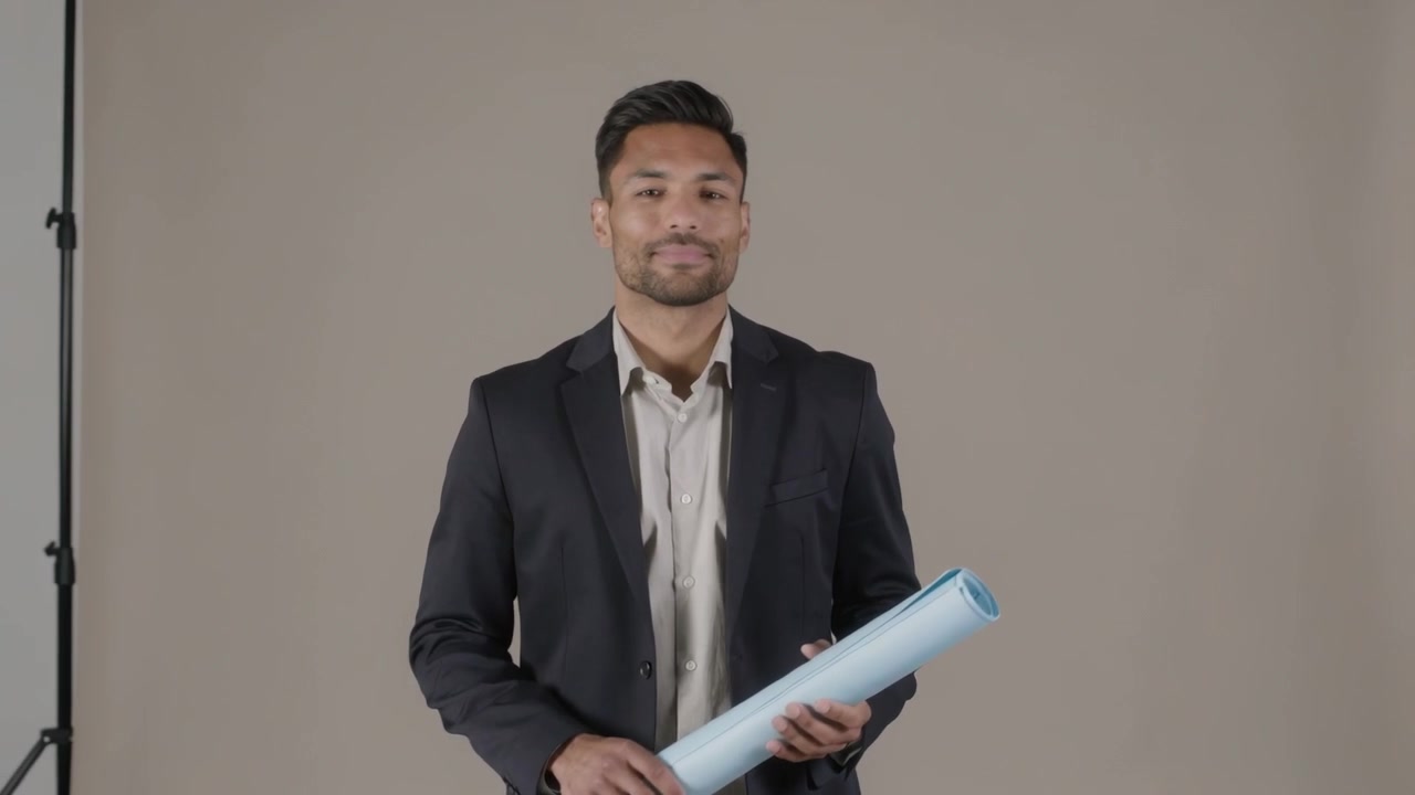 Grabación comenzando, hombre con blazer presentando mientras cambia de pose y sostiene un tubo de planos azul en un estudio