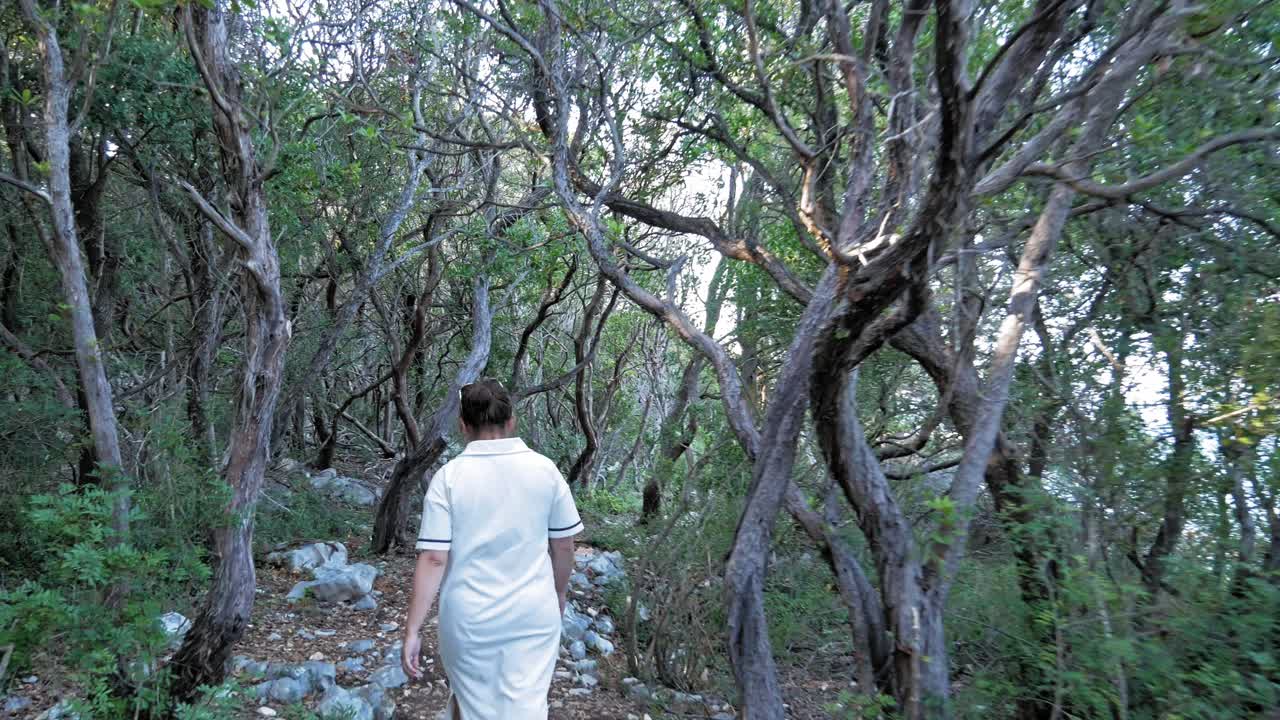 vista trasera de una mujer vestida de blanco caminando por el bosque hacia la playa emplisi en grecia