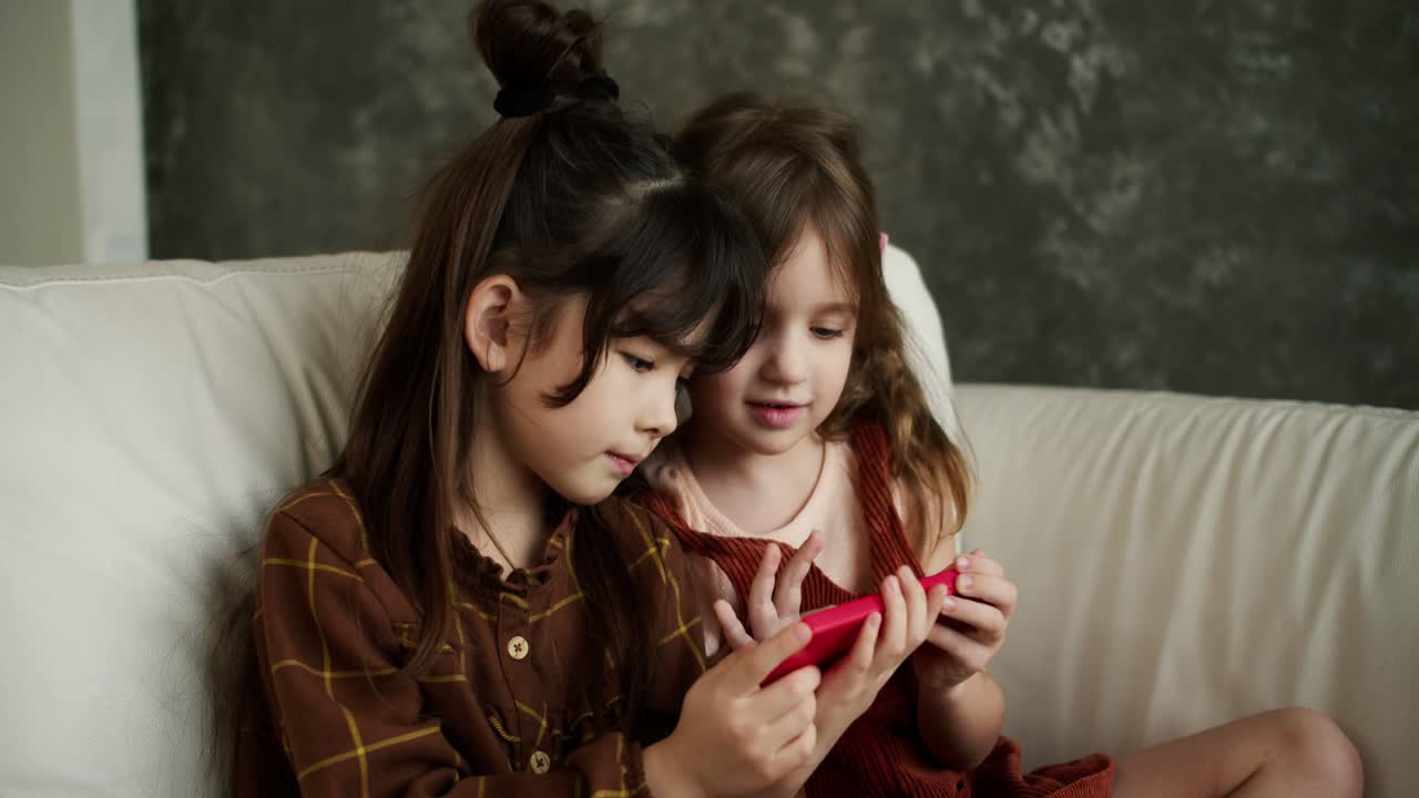 Two smiling children sit on couch look at cellphone screen, have fun together