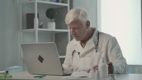 a grey haired, middle aged doctor consults on his laptop the composition of a medicine 1