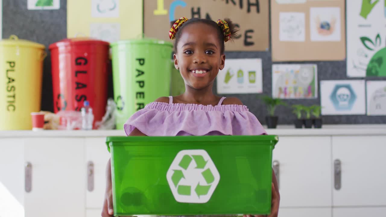 feliz colegiala afroamericana de pie en el aula, sonriendo, sosteniendo un cubo de reciclaje