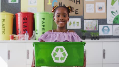 Happy-african-american-schoolgirl-standing-in-classroom,-smiling,-holding-recycling-bin