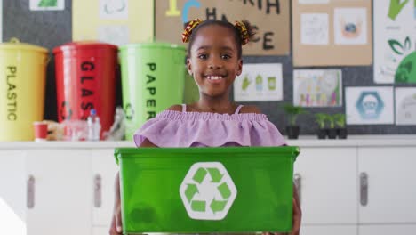 happy african american schoolgirl standing in classroom, smiling, holding recycling bin