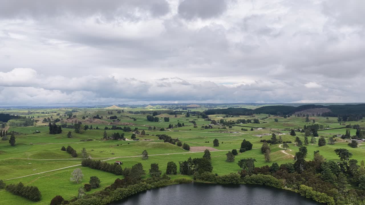 Luftoptagelse med indzoom over en rolig sø mod bølgende grønt landbrugsland i New Zealand, droneudsigt over landbrugsjord og filmisk landskab.