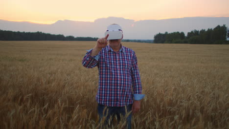 an elderly male farmer uses gestures with his hands standing in a field wearing virtual reality glasses. use vr glasses in agriculture on a field with wheat.