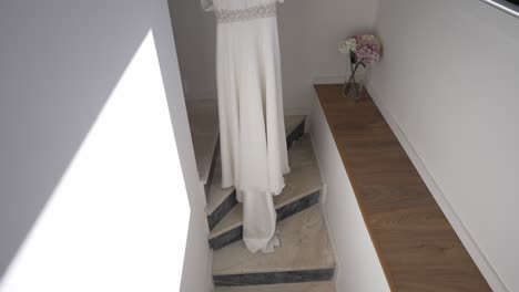 Weißes-Brautkleid-Hängt-An-Einer-Treppe,-Sonnenlicht-Wirft-Schatten,-Blumen-Auf-Einem-Beistelltisch