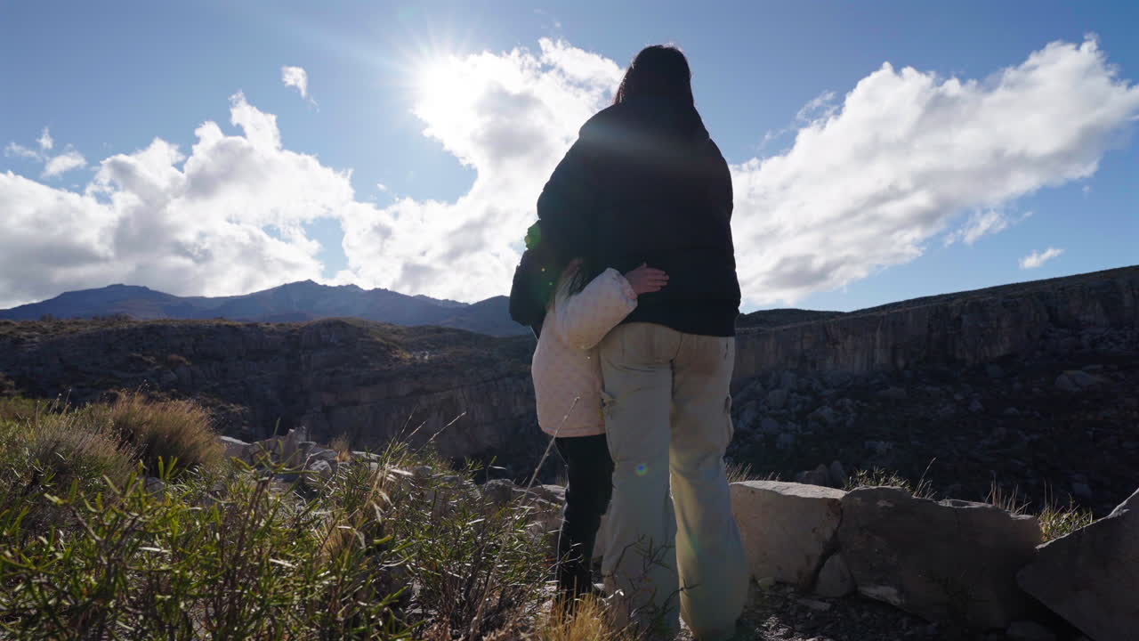 Mother and daughter hugging while admiring mountain landscape under sunlight, family love and travel