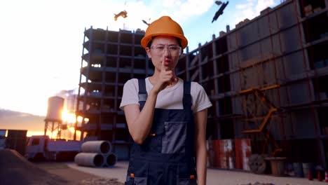 trabajadora asiática con gafas de protección y casco de seguridad mirando a la cámara y haciendo un gesto de silencio mientras está de pie en el sitio de construcción