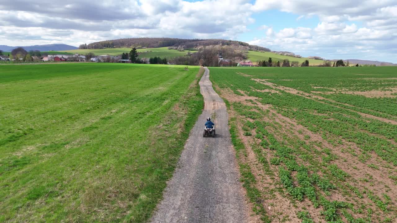 Drone che segue un bambino in quad lungo una lunga strada di campagna attraverso aperti campi verdi, ampia vista aerea che mostra una campagna serena e colline lontane
