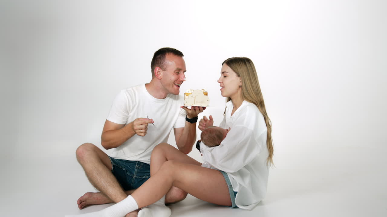 Parents take a bite from a cake, laugh and kiss. Happy family celebrating the first month of their baby.
