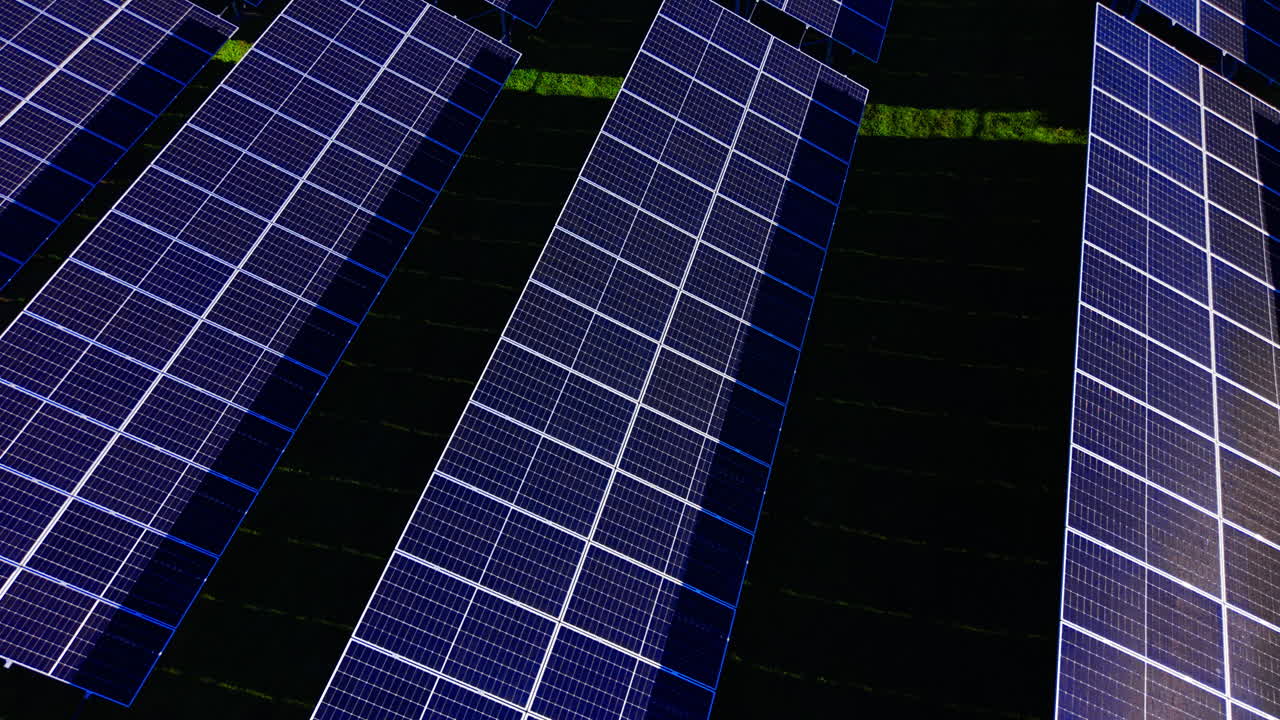 Vista dall'alto di moduli solari che mostrano un riflesso solare luminoso sulla superficie del pannello, in contrasto con il terreno scuro e l'erba verde. Vista aerea