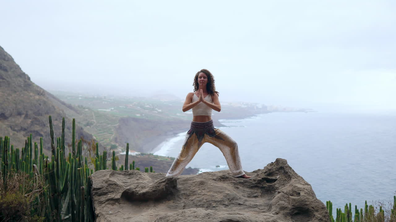 posicionada por el océano, la playa y las montañas rocosas, una mujer medita en la postura del guerrero del yoga, canalizando la motivación y la inspiración en su rutina de fitness