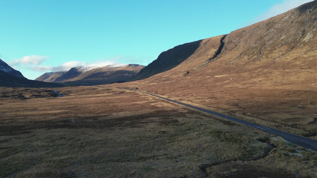 Carretera remota en el valle serpenteando por Glencoe con imponentes montañas, cielos despejados, Escocia, vista aérea.