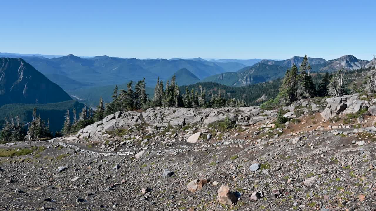 Cinematic aerial view over rocky alpine terrain with distant blue mountains and sparse pine trees under a clear sky