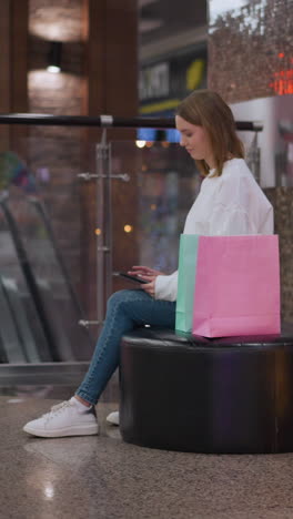 mujer joven sentada en un banco circular en un centro comercial usando su teléfono, rodeada de bolsas de compras, estrella iluminada decorativa y pantalla de luz brillante