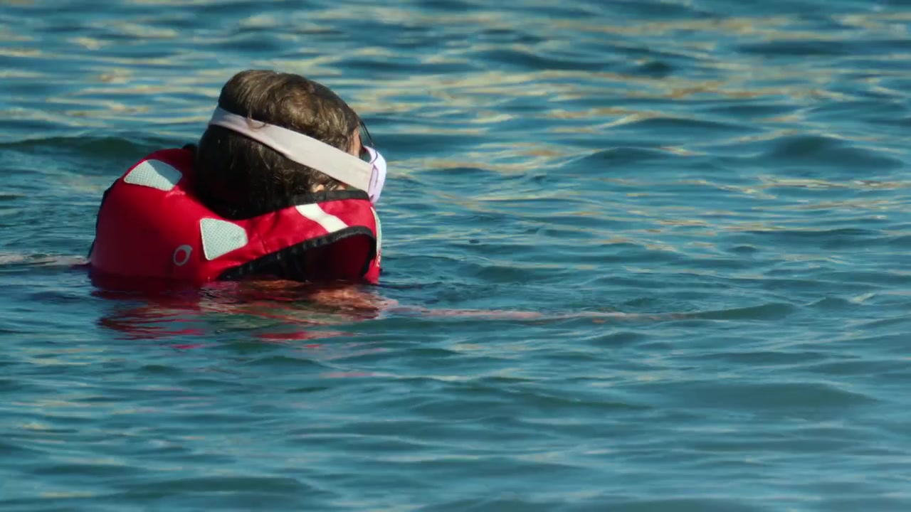 Bambino che indossa un giubbotto di salvataggio rosso e occhialini mentre nuota in acque blu e calme, concentrandosi sulla sicurezza in acqua e sul divertimento.