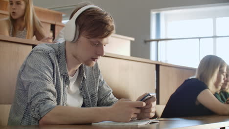 A-student-sitting-in-a-university-auditorium-with-a-large-group-of-people-sits-in-headphones-and-listens-to-music-and-looks-at-the-smartphone-screen.-It-is-in-a-state-of-calm.-Class-break