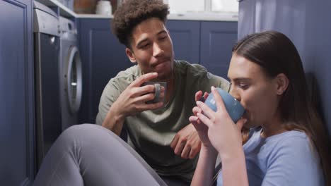 Happy-biracial-couple-sitting-on-floor-in-kitchen,-drinking-coffee